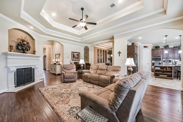 2103 Matagorda Ln, Friendswood, TX 77546 - living room featuring arched walkways, a ceiling fan, a raised ceiling, a fireplace with raised hearth, and light wood-type flooring
