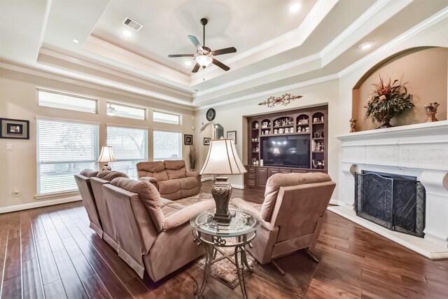 living room with a raised ceiling, a ceiling fan, dark wood finished floors, a fireplace with raised hearth, and ornamental molding