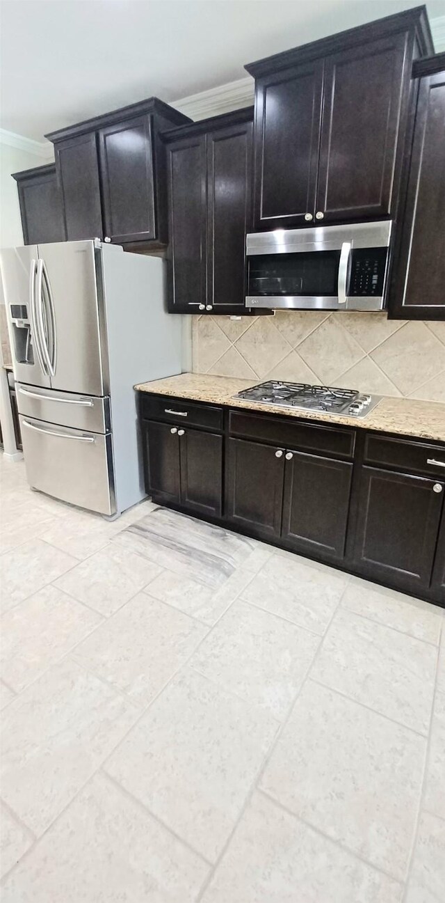kitchen featuring stainless steel appliances, crown molding, backsplash, dark cabinets, and light stone counters