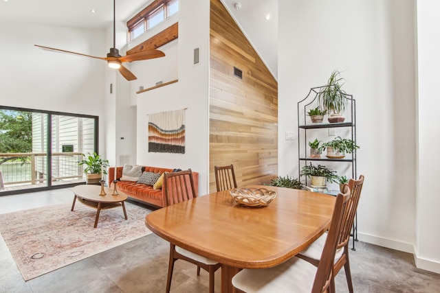 dining room featuring a towering ceiling, finished concrete floors, a ceiling fan, wood walls, and baseboards