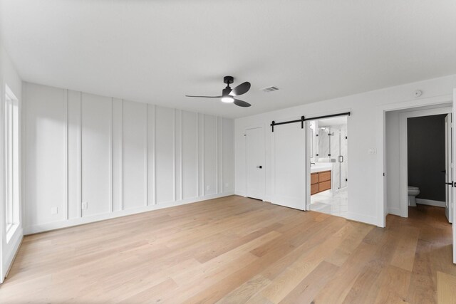 unfurnished bedroom featuring a barn door, light wood finished floors, ensuite bath, a decorative wall, and a ceiling fan