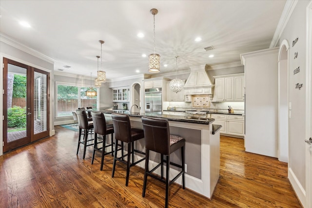 kitchen featuring custom range hood, stainless steel built in fridge, ornamental molding, a kitchen bar, and a large island with sink