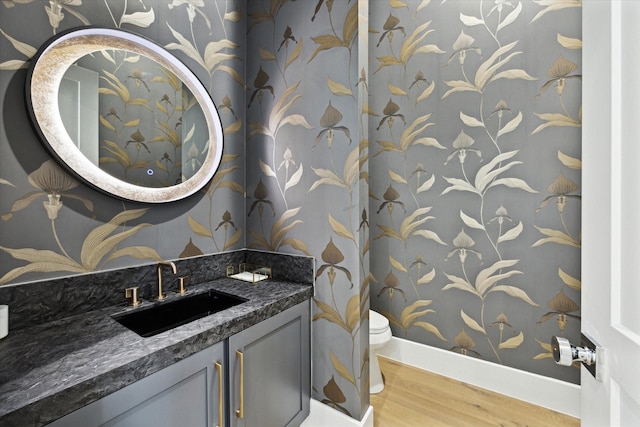 half bathroom featuring wood finished floors, toilet, wallpapered walls, vanity, and baseboards