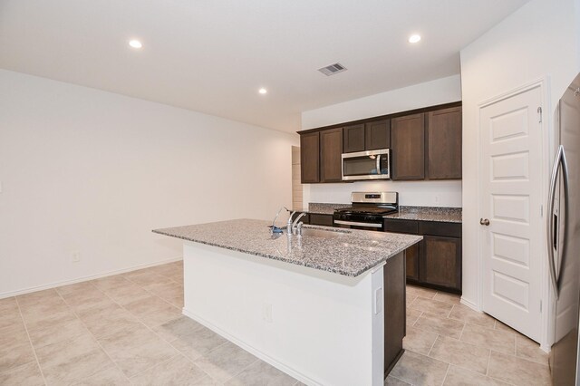 kitchen with appliances with stainless steel finishes, an island with sink, dark brown cabinets, light stone countertops, and recessed lighting