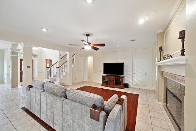 living area featuring ceiling fan, a tiled fireplace, light tile patterned floors, ornamental molding, and stairs