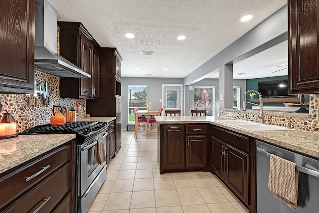 kitchen featuring wall chimney range hood, appliances with stainless steel finishes, a sink, light tile patterned floors, and dark brown cabinets