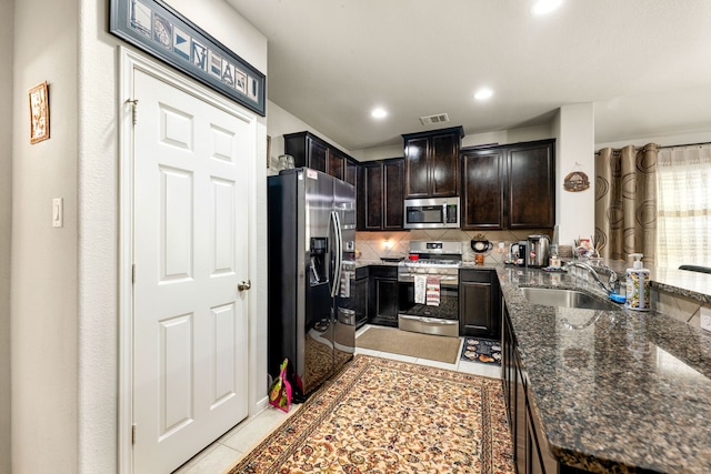 kitchen featuring appliances with stainless steel finishes, a sink, decorative backsplash, dark stone countertops, and dark brown cabinetry