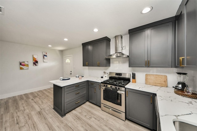 kitchen with wall chimney range hood, stainless steel range with gas stovetop, a peninsula, light stone countertops, and baseboards