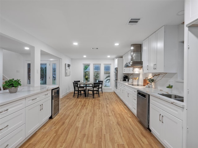 kitchen with wall chimney exhaust hood, wine cooler, appliances with stainless steel finishes, light wood-type flooring, and recessed lighting