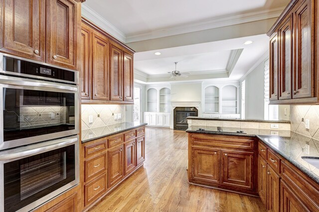 kitchen with double oven, ceiling fan, a glass covered fireplace, dark stone countertops, and light wood finished floors