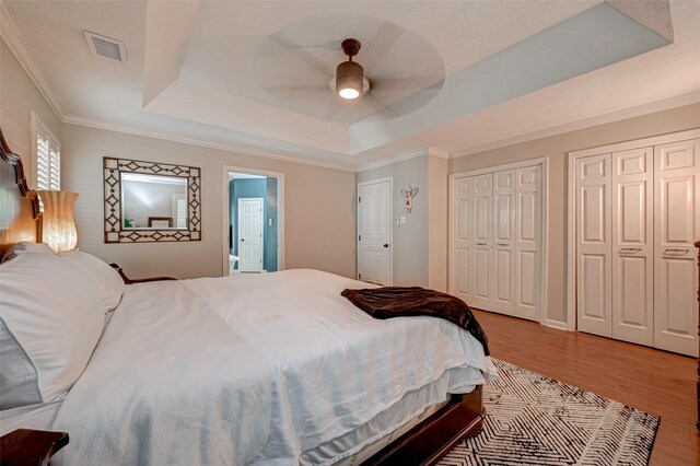bedroom with a raised ceiling, multiple closets, wood finished floors, crown molding, and ceiling fan