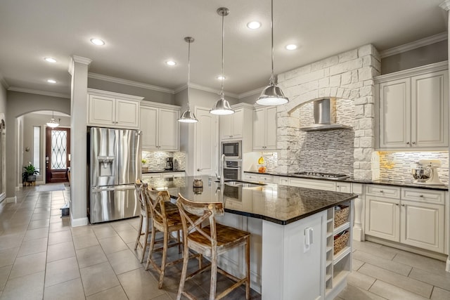 kitchen with stainless steel appliances, wall chimney range hood, arched walkways, dark countertops, and open shelves