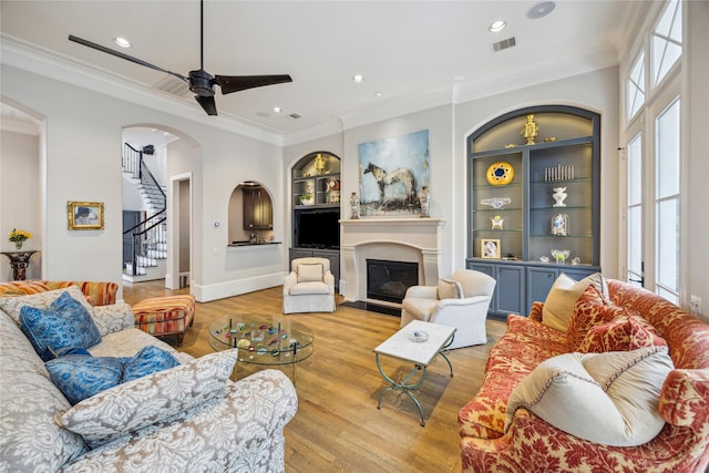 living area with built in shelves, wood finished floors, crown molding, stairs, and arched walkways
