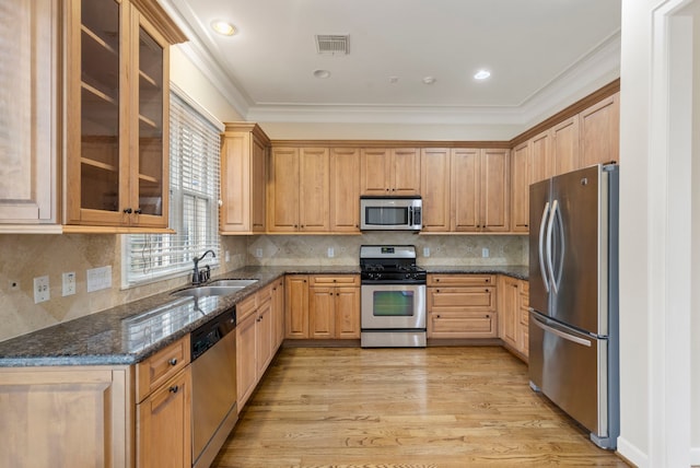 kitchen featuring appliances with stainless steel finishes, a sink, dark stone counters, light wood finished floors, and glass insert cabinets