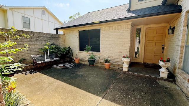 property entrance featuring roof with shingles, brick siding, a patio, and fence