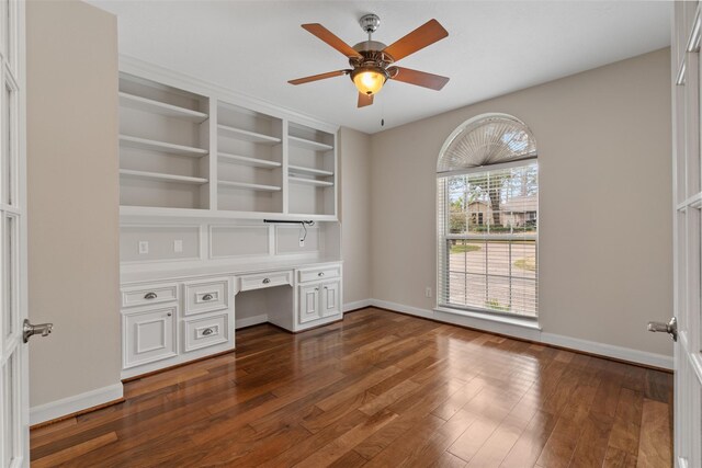 unfurnished office with built in desk, baseboards, dark wood finished floors, and a ceiling fan