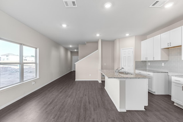 kitchen featuring backsplash, dark wood-style floors, a kitchen island with sink, light stone counters, and baseboards