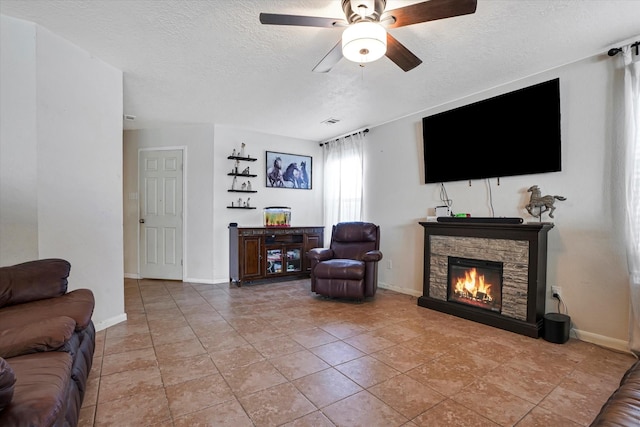 living room with a ceiling fan, a textured ceiling, a stone fireplace, baseboards, and light tile patterned flooring