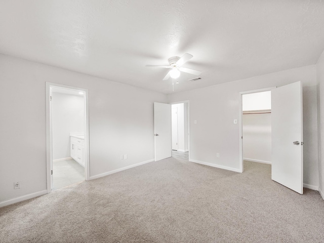unfurnished bedroom with light colored carpet, a walk in closet, baseboards, ensuite bathroom, and ceiling fan