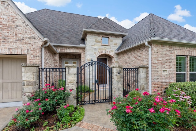 view of exterior entry with a gate, brick siding, a shingled roof, and fence