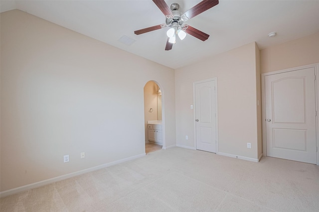 unfurnished bedroom featuring light carpet, arched walkways, baseboards, ensuite bathroom, and a ceiling fan