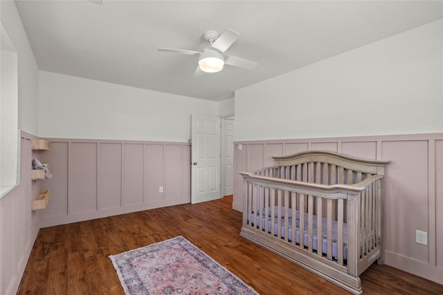 bedroom featuring a decorative wall, wainscoting, wood finished floors, a ceiling fan, and a nursery area