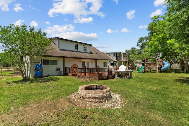 rear view of property featuring a fire pit, a wooden deck, a lawn, a playground, and roof with shingles