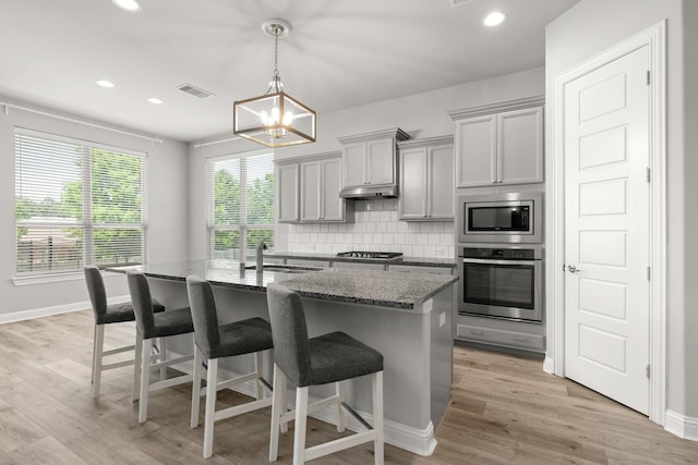 kitchen featuring stainless steel appliances, a sink, dark stone counters, an island with sink, and backsplash