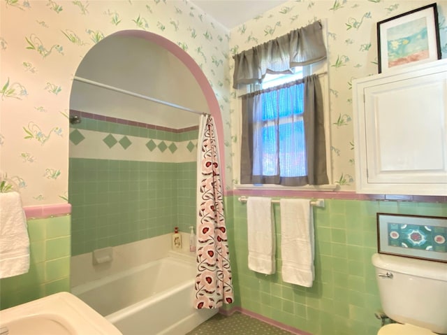 full bathroom featuring wallpapered walls, wainscoting, tile walls, shower / bathtub combination with curtain, and toilet
