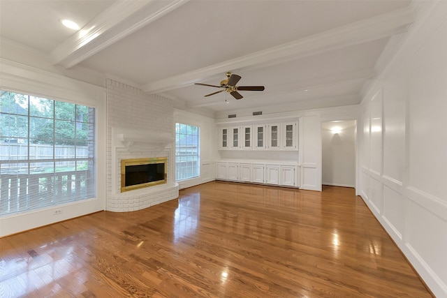 unfurnished living room featuring beamed ceiling, wood finished floors, a ceiling fan, a brick fireplace, and crown molding