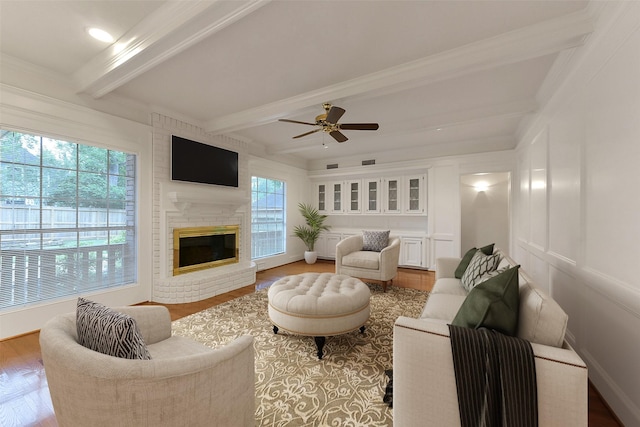 living area featuring beam ceiling, wood finished floors, a fireplace, a ceiling fan, and ornamental molding