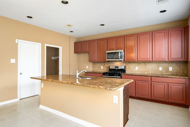 kitchen featuring appliances with stainless steel finishes, a sink, decorative backsplash, light stone counters, and an island with sink