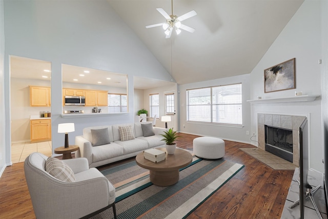 living room featuring high vaulted ceiling, baseboards, light wood finished floors, a ceiling fan, and a tile fireplace