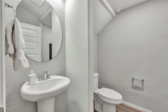 half bath featuring wood finished floors, toilet, a textured wall, a sink, and lofted ceiling