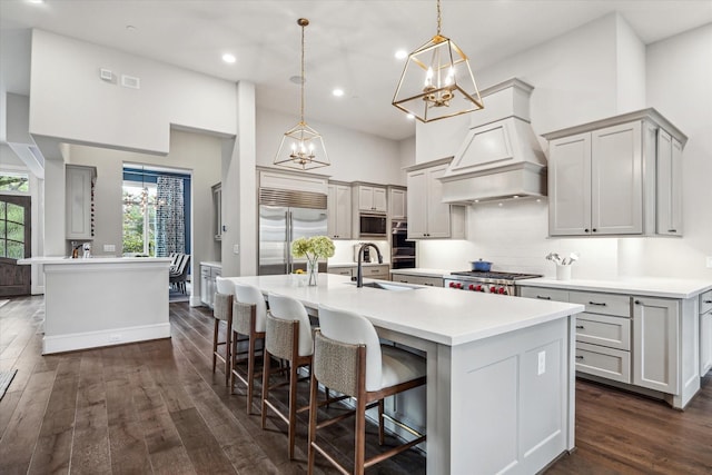 kitchen featuring stainless steel appliances, a chandelier, a sink, a kitchen island with sink, and premium range hood