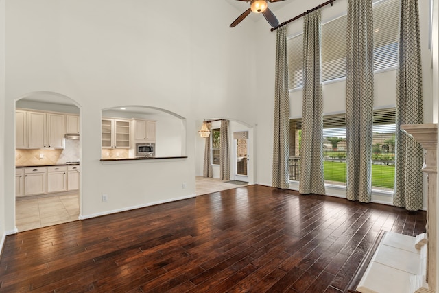 unfurnished living room featuring a high ceiling, ceiling fan, light wood finished floors, arched walkways, and baseboards