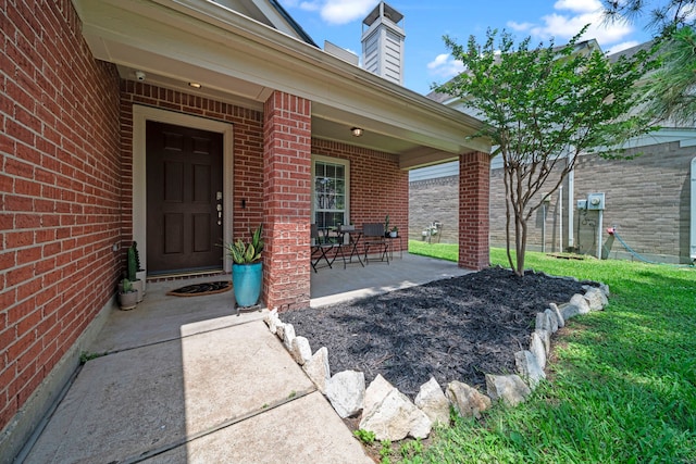 entrance to property featuring brick siding, a chimney, a yard, and covered porch