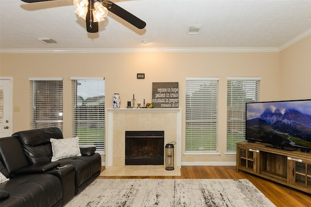 living area featuring wood finished floors, a ceiling fan, crown molding, a tiled fireplace, and baseboards
