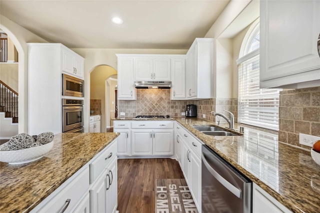 kitchen with appliances with stainless steel finishes, a sink, arched walkways, under cabinet range hood, and white cabinets