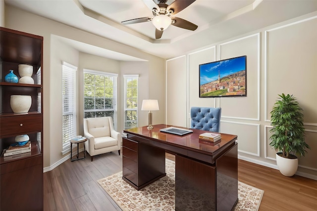 office space featuring a ceiling fan, a decorative wall, a tray ceiling, and wood finished floors