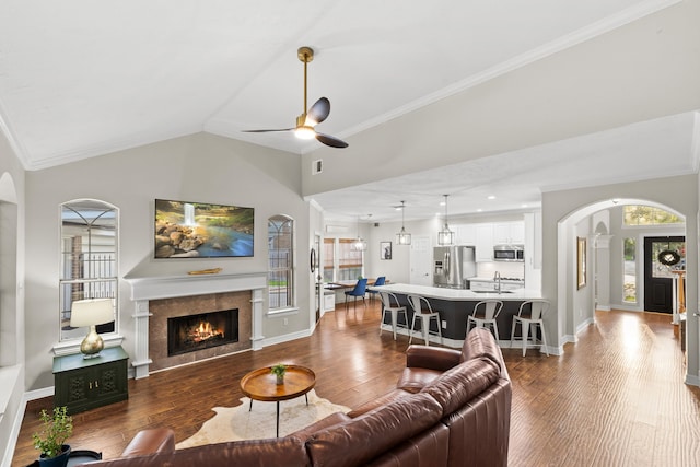 living room featuring arched walkways, a warm lit fireplace, ornamental molding, a ceiling fan, and wood finished floors