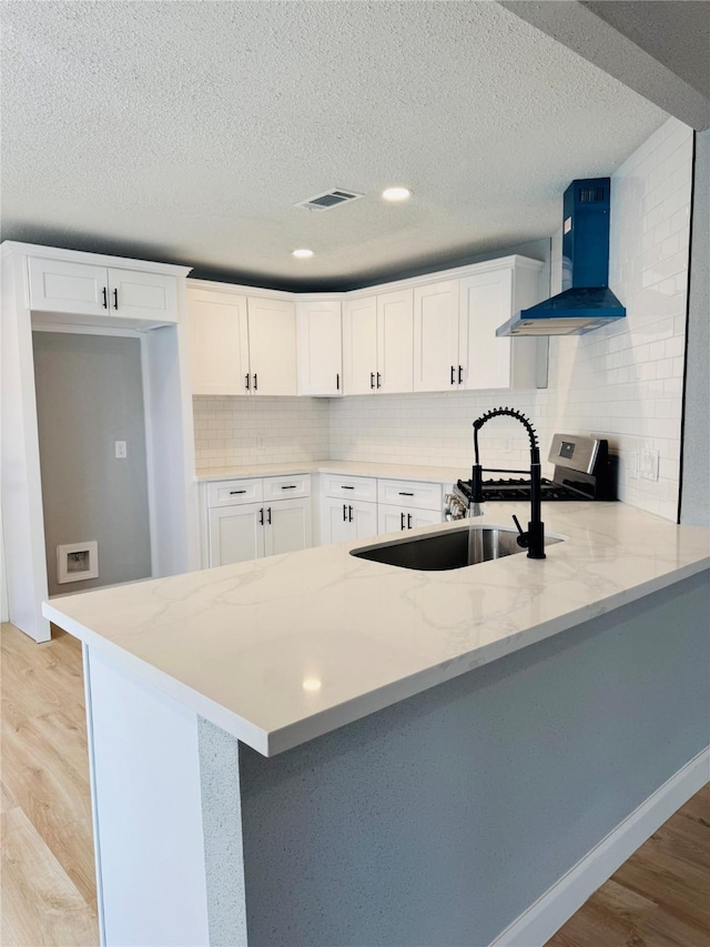 kitchen featuring wall chimney exhaust hood, a peninsula, light wood-style floors, white cabinets, and a textured ceiling