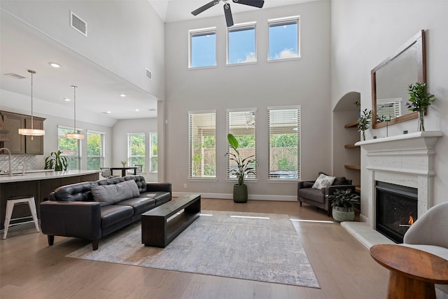 living room featuring ceiling fan, a fireplace, light wood-style flooring, a towering ceiling, and baseboards