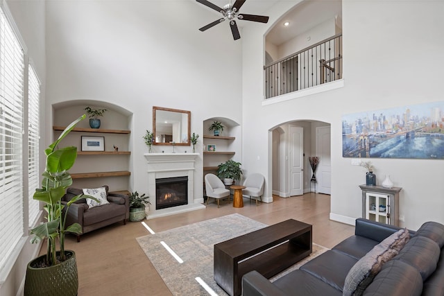 living area featuring built in shelves, arched walkways, a glass covered fireplace, wood finished floors, and a ceiling fan