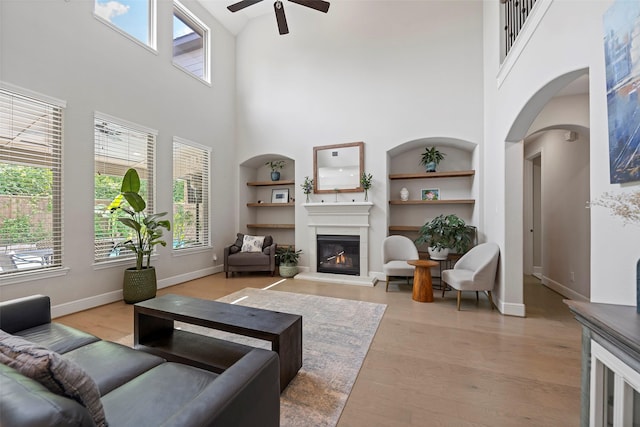 living room with ceiling fan, a towering ceiling, built in shelves, a glass covered fireplace, and baseboards