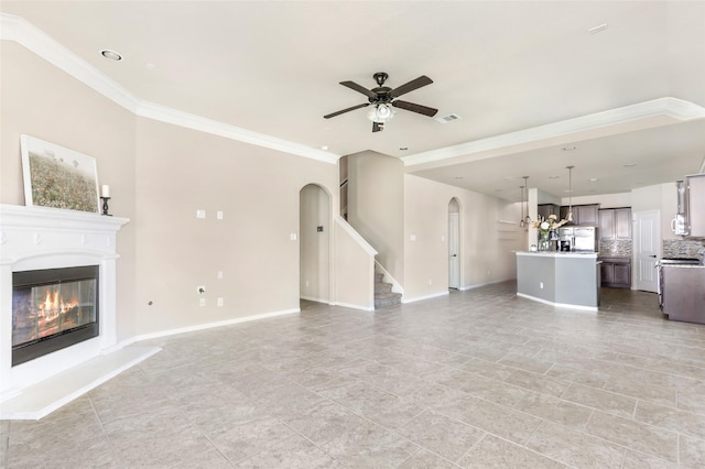 unfurnished living room featuring arched walkways, a glass covered fireplace, a ceiling fan, crown molding, and stairway