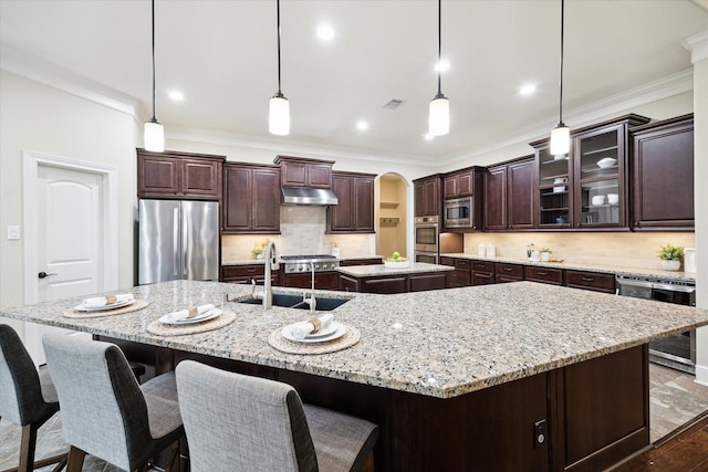 kitchen with stainless steel appliances, a sink, a large island with sink, under cabinet range hood, and crown molding