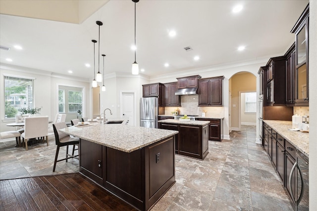 kitchen with a large island with sink, freestanding refrigerator, under cabinet range hood, ornamental molding, and arched walkways