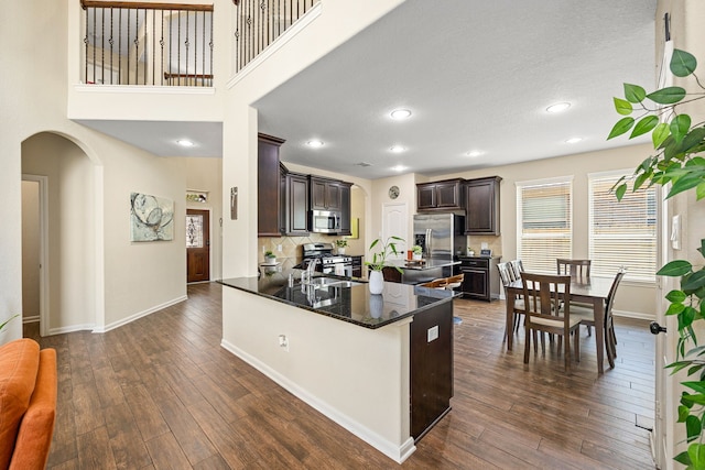 kitchen with arched walkways, appliances with stainless steel finishes, dark brown cabinetry, dark wood-style flooring, and baseboards