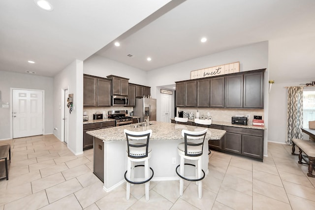 4827 San Gregorio Ln, Katy, TX 77493 - kitchen featuring appliances with stainless steel finishes, dark brown cabinets, a kitchen island with sink, decorative backsplash, and light stone countertops
